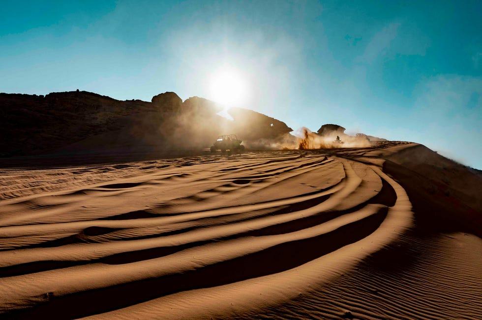 une photo éloignée d'amateurs d'aventure conduisant un buggy tout-terrain alors que le soleil couchant met en évidence les traces du buggy et les sables sculptés par le vent d'Al Ula